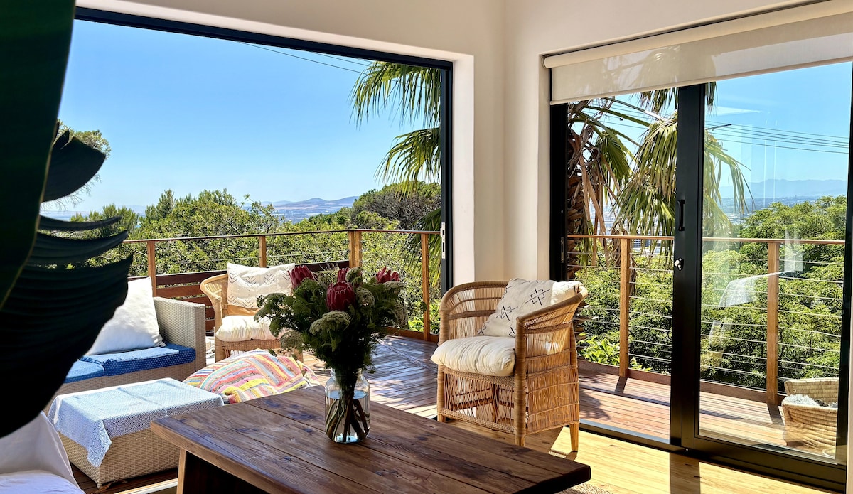 A spacious balcony is visible, featuring a wooden table surrounded by wicker seating. Lush greenery is present in the background, framed by glass sliding doors that open to stunning views of the landscape. Sunlight bathes the area, creating a bright and inviting atmosphere.