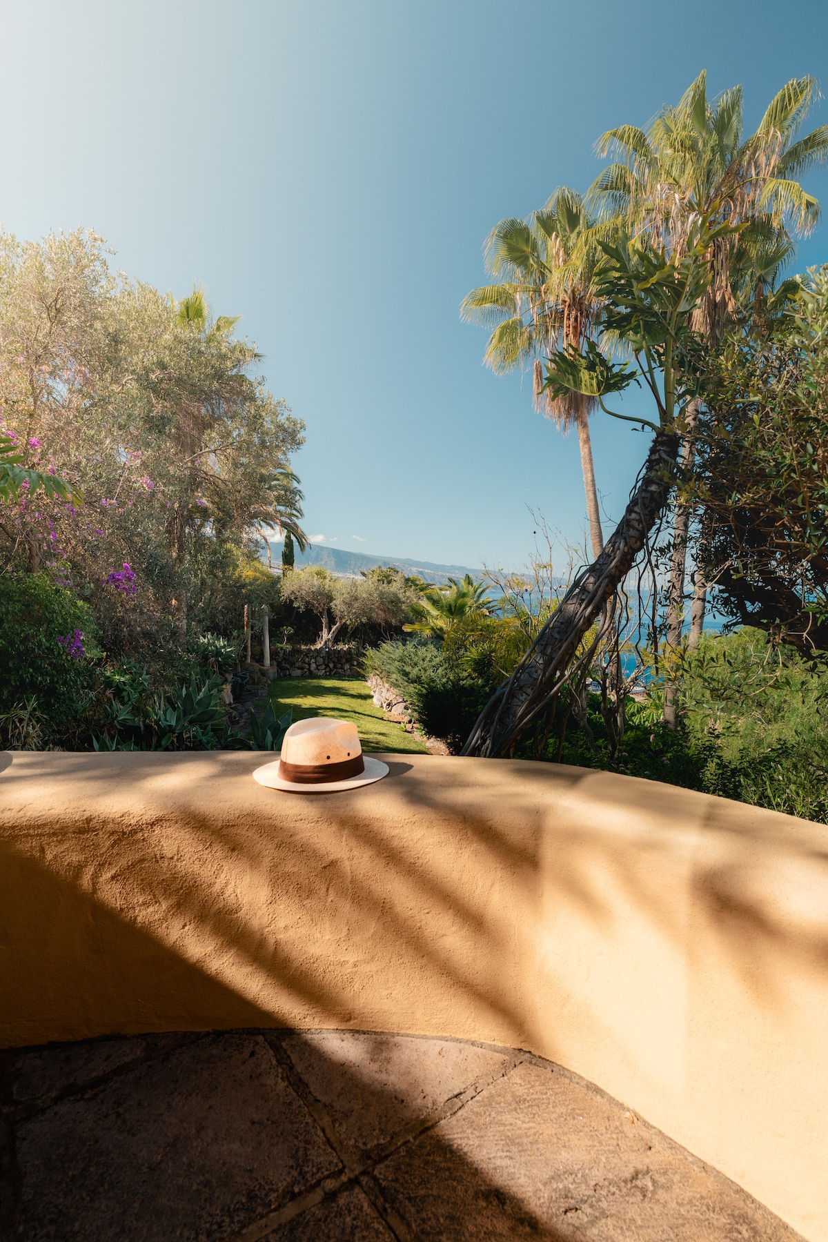 A sunlit terrace features a wide stone ledge with a straw hat placed atop it. Lush greenery surrounds the area, including palm trees and flowering plants, while the expansive blue sky and distant ocean create a serene backdrop.