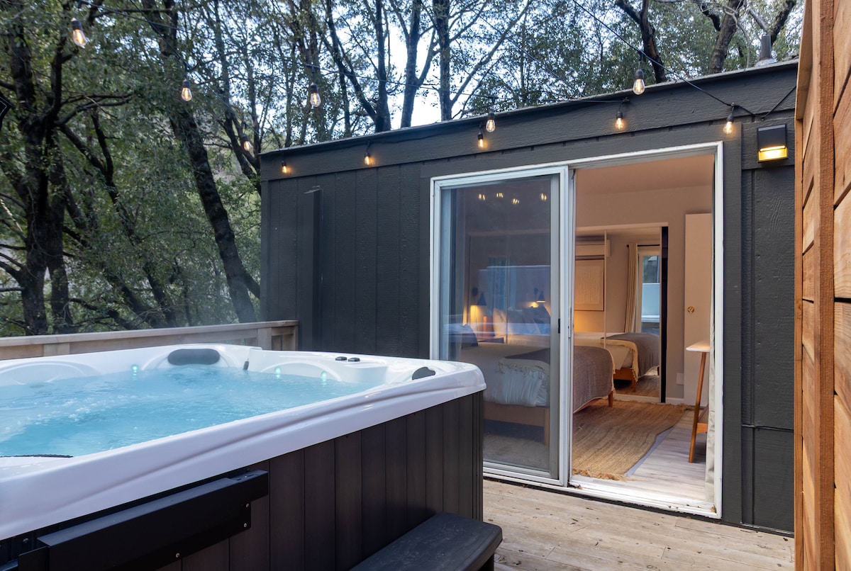 A private hot tub is positioned on a wooden deck, surrounded by trees. Soft lighting illuminates the area, creating a serene ambiance. The sliding glass doors reveal an interior view of the cabin's bedrooms, enhancing the connection between indoor and outdoor relaxation spaces.