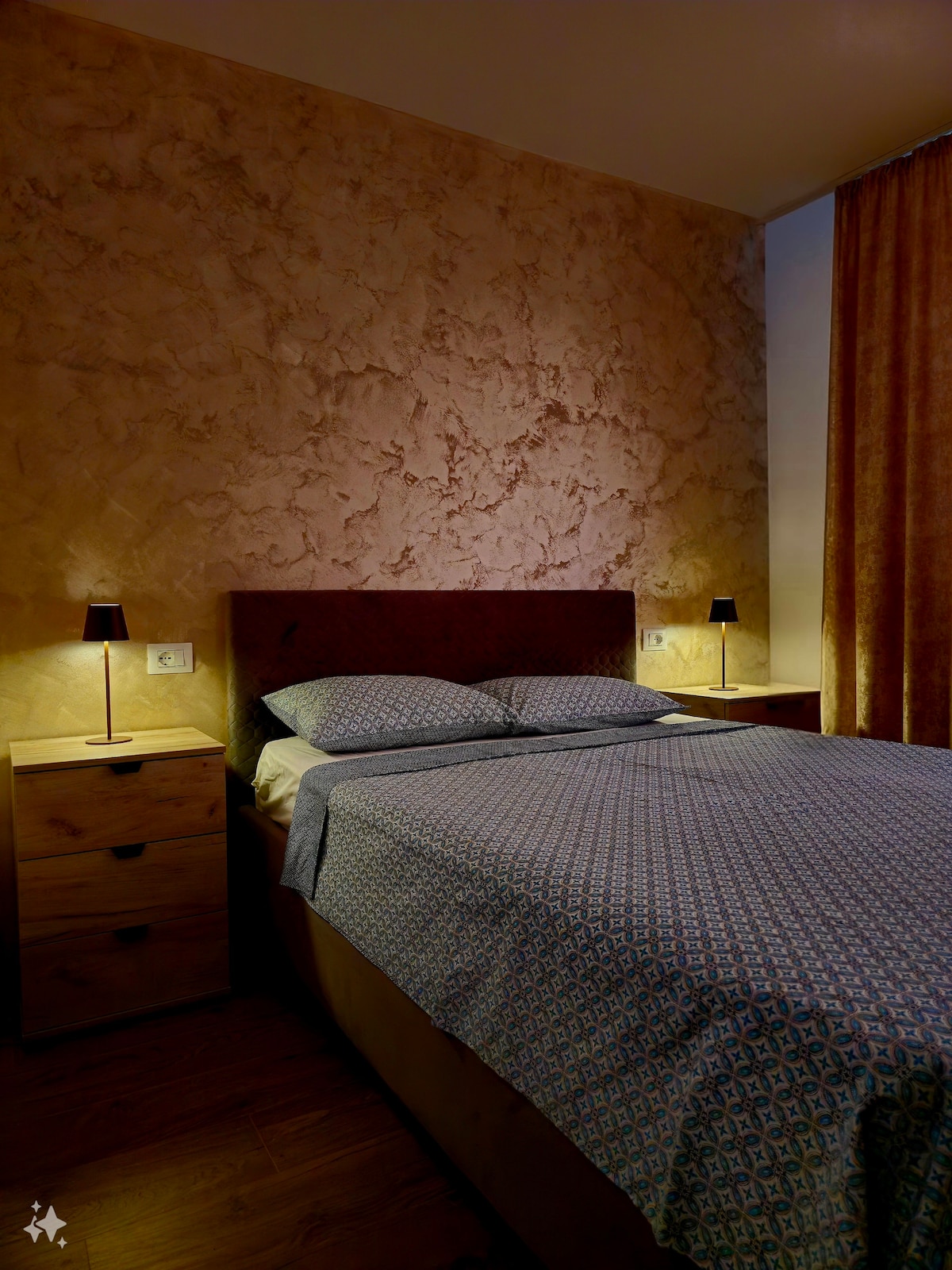 A cozy bedroom is presented with a spacious bed dressed in a patterned blanket. Two bedside tables stand on either side, each featuring a small lamp. The warm, textured wall creates a calm ambiance, complemented by soft natural light filtering through the curtains.