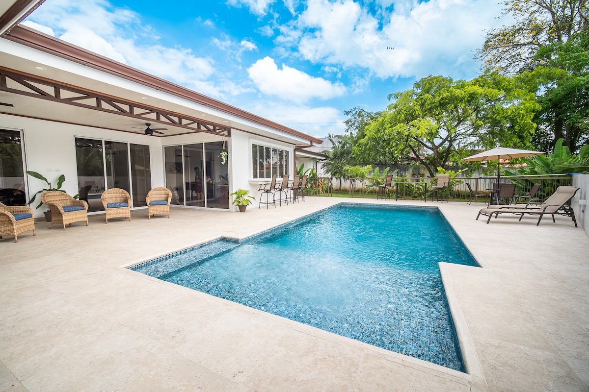 A spacious outdoor patio features a refreshing swimming pool, surrounded by loungers and tropical plants. Sunlight reflects off the water, creating a serene atmosphere, while large glass doors offer a view into the adjoining living area.
