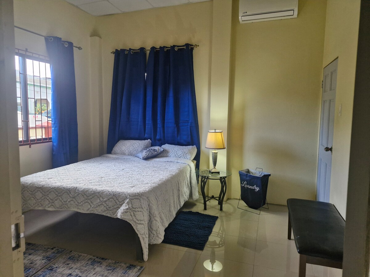 A spacious bedroom features a large bed with a light-colored patterned comforter. A bedside table with a lamp is positioned next to the bed, while dark curtains frame the window. A small bench and a laundry basket are visible, adding to the room's functionality.
