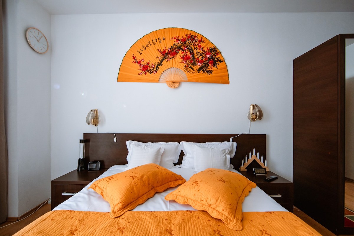 A bedroom featuring a spacious bed adorned with vibrant orange pillows and a coordinating blanket. An art piece resembling a fan hangs above the bed, adding a decorative touch. Table lamps are positioned on each side, complemented by a wooden nightstand.
