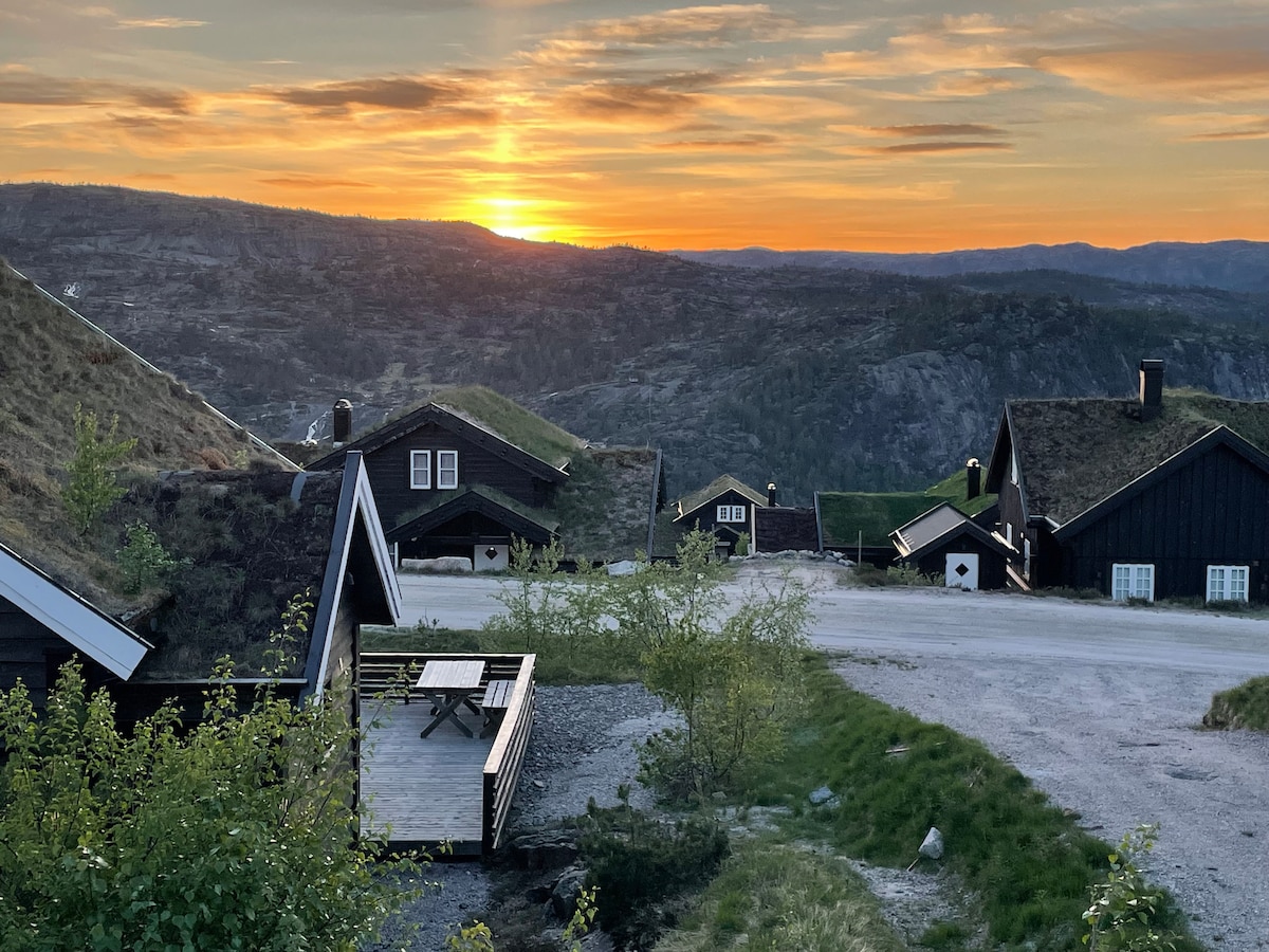 A serene landscape is captured at sunset, with the sun casting warm tones over rolling hills. Several cabins with green roofs are positioned along a gravel pathway. A wooden deck with seating is visible, surrounded by lush greenery, enhancing the rustic charm of the location.