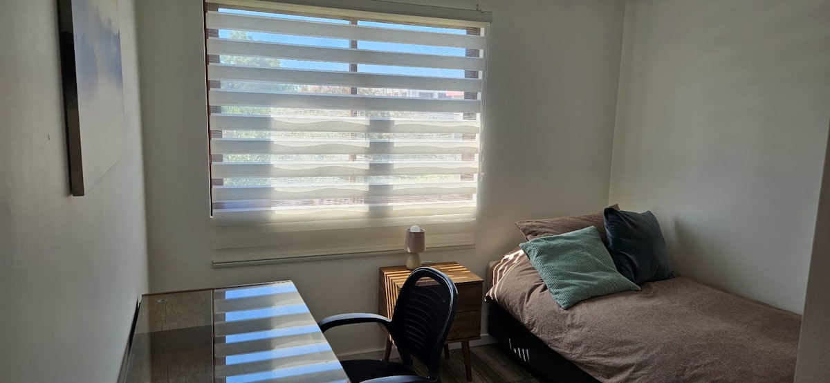 A bright sleeping area features a spacious bed dressed in soft linens, accompanied by decorative pillows. A modern desk with a chair is positioned nearby, and natural light filters through window blinds, creating a calm atmosphere.