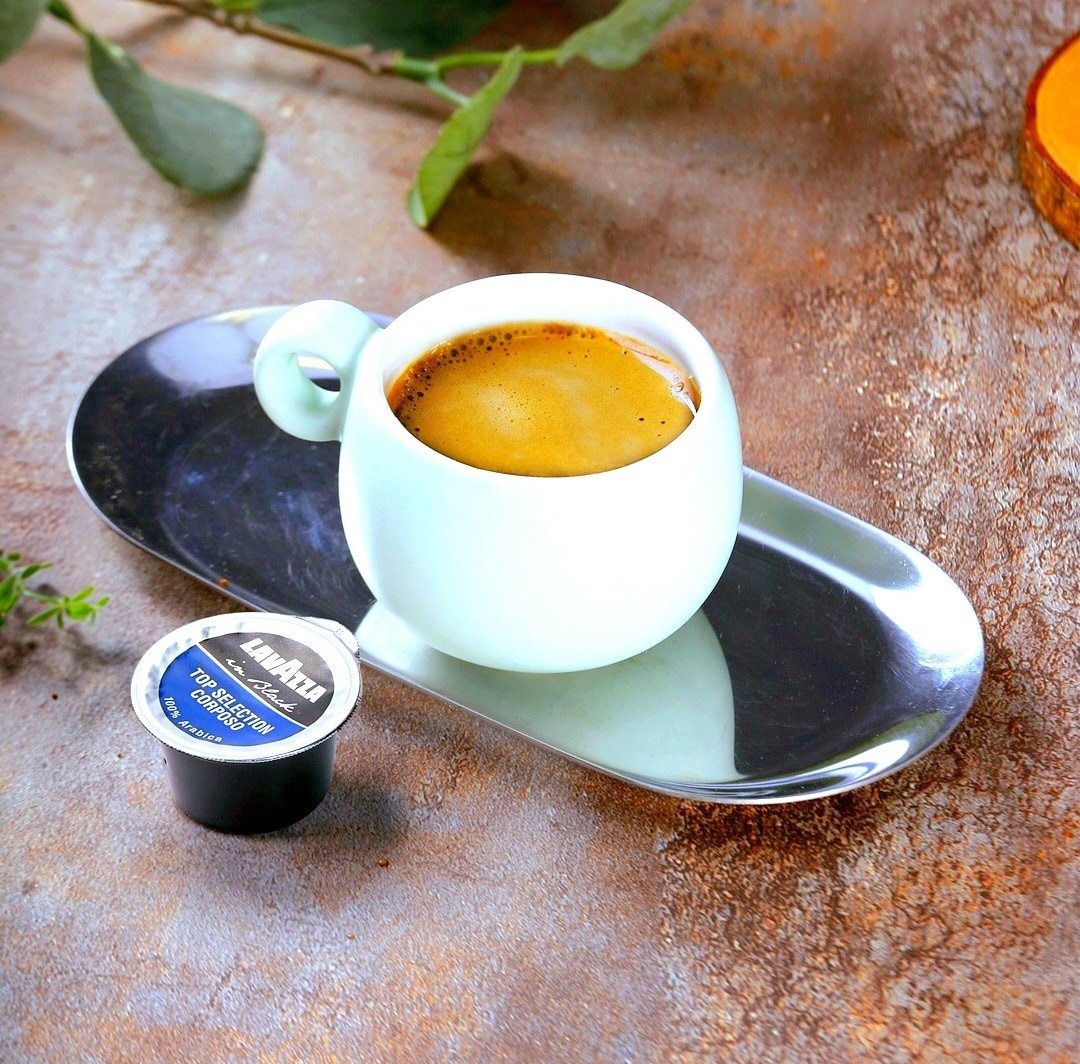 A white coffee cup contains a rich, dark espresso, resting on a sleek gray plate. Next to the cup, a Lavazza coffee capsule is displayed. The earthy backdrop suggests a warm and inviting coffee experience.