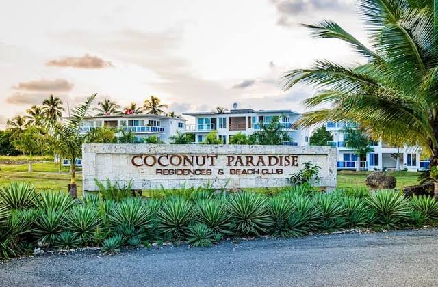 The entrance to Coconut Paradise Beach Club is marked by a stone sign featuring the name prominently displayed. Lush greenery and palm trees surround the area, evoking a tranquil atmosphere, while the light of dusk casts a warm glow over the scene.