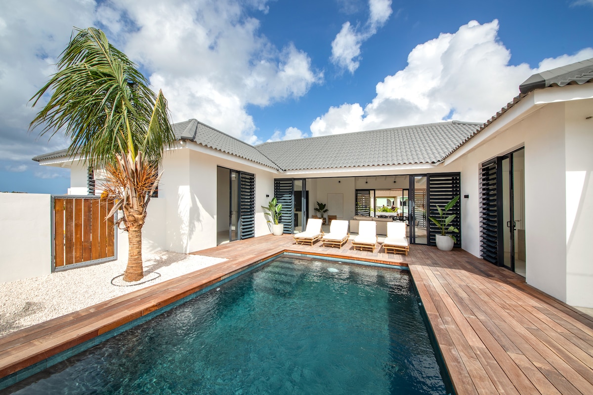 A serene outdoor area is highlighted by a Balinese-style pool surrounded by a wooden deck. Lounge chairs are positioned near the water, offering a place to relax. Lush greenery and palm trees are present, complemented by a bright blue sky with scattered clouds.