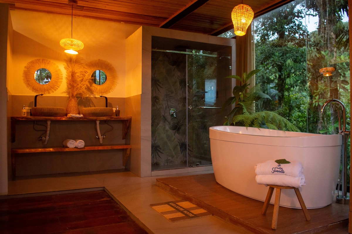 A spa-like bathroom features a freestanding tub positioned near a large window, offering views of lush greenery. Two circular mirrors are mounted above a wooden vanity that holds complimentary toiletries. Soft lighting from woven pendant fixtures creates a relaxed ambiance.