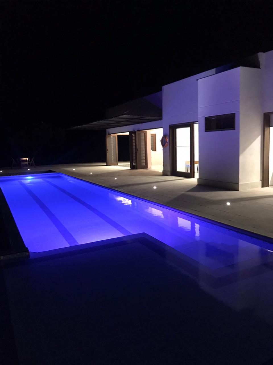 A well-lit pool area is shown at night, where the water is illuminated in calming blue tones. The modern structure nearby features large glass doors, providing a view of the interior. Subtle lighting enhances the surrounding patio space.