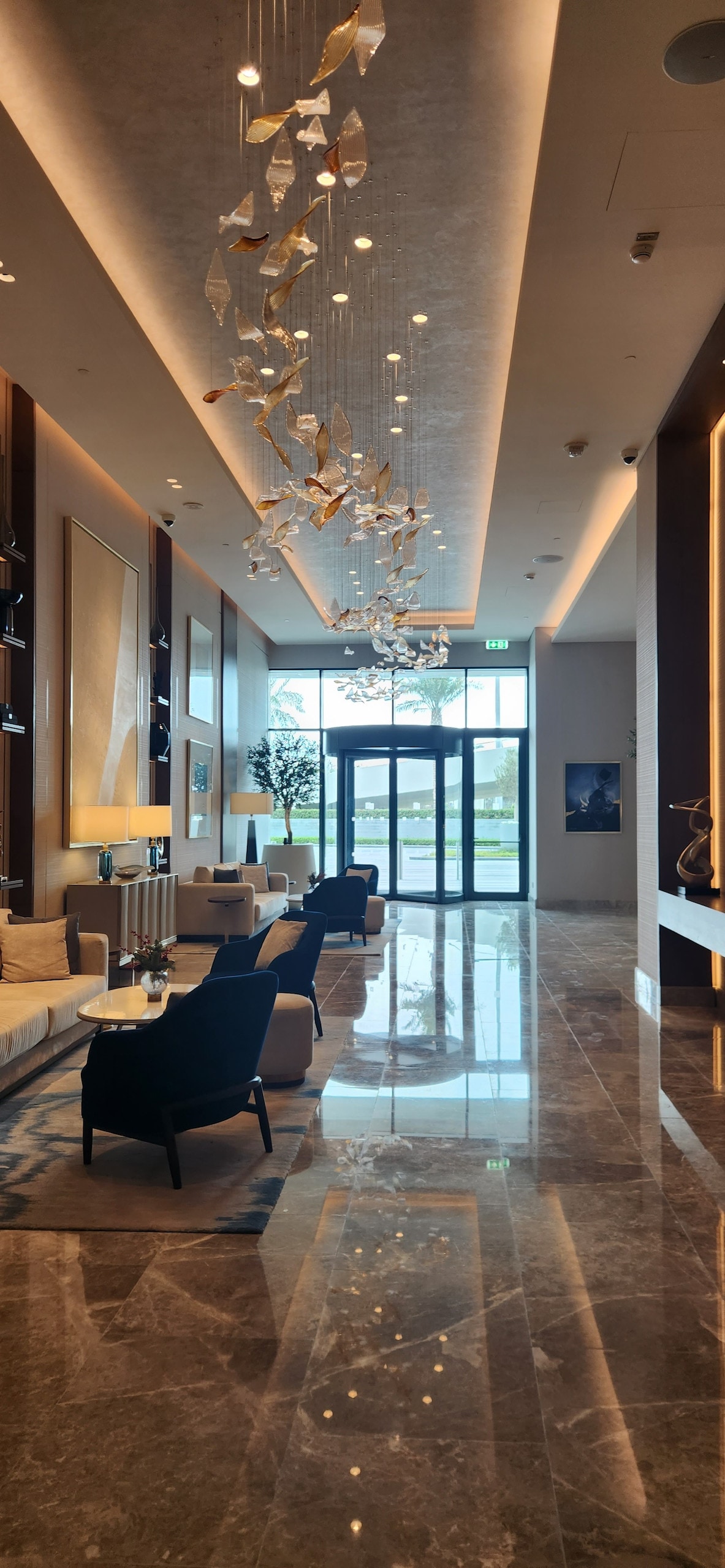 A modern lobby features polished marble flooring, with a spacious seating area that includes several chairs and small tables. Natural light filters through large glass doors at the entrance, and decorative art pieces adorn the walls alongside an artistic light fixture overhead.