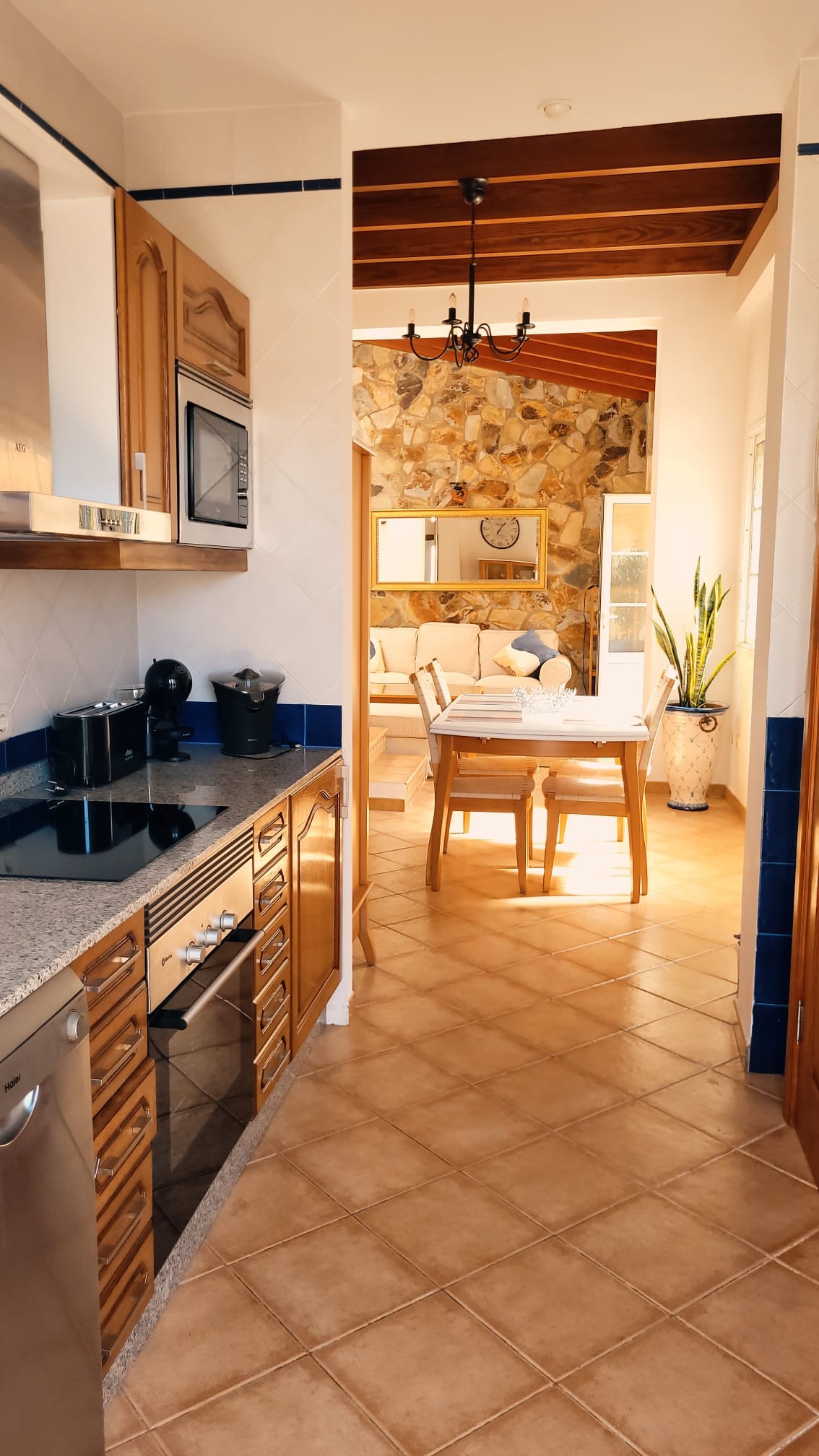 A well-designed kitchen occupies one side of the space, featuring wooden cabinetry and modern appliances. Natural light enhances the inviting atmosphere, leading to a dining area with a table and chairs. An interesting stone wall adds character, complemented by greenery near the windows.