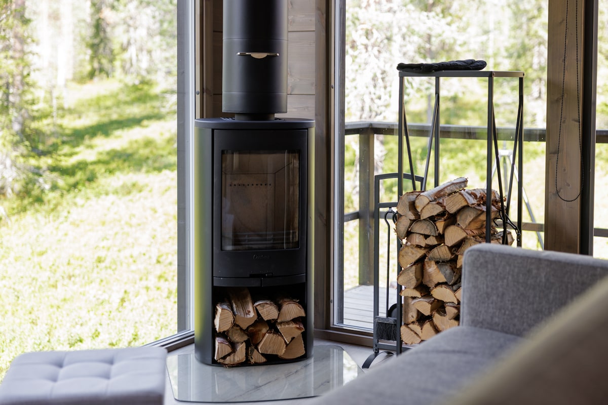 A black wood-burning stove is positioned next to a stack of firewood, providing warmth and comfort. Large windows allow for a view of the serene forest landscape outside, while a cozy sofa invites relaxation nearby.
