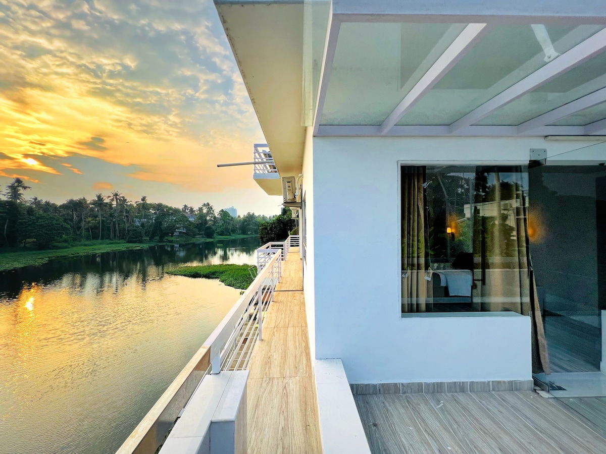 A spacious balcony overlooks the tranquil waters of the Muttar River, reflecting the soft hues of sunrise. The nearby riverbank is framed by lush greenery, enhancing the peaceful setting. A large window allows natural light to fill the interior space, creating a serene ambiance.