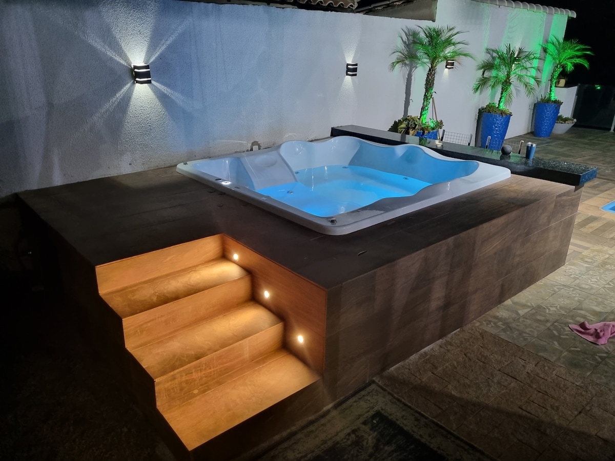 A well-lit jacuzzi area is shown at night, featuring soft blue water. Wooden steps lead up to the jacuzzi, with recessed lighting illuminating the path. Potted plants are positioned throughout the area, adding a touch of greenery in the background.