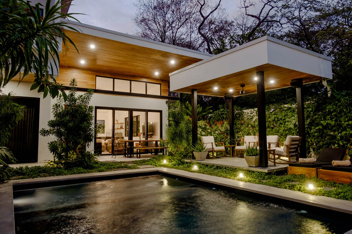 The exterior of a modern house is showcased, featuring a private pool surrounded by lush greenery. A covered patio with seating areas is visible, along with large glass doors that open into the main living space, creating a connection with the outdoors.