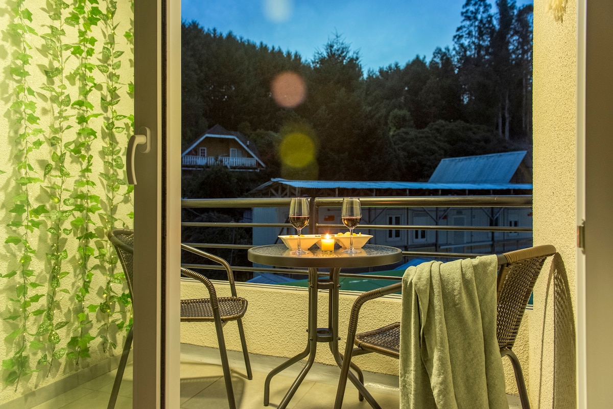 A private balcony is equipped with a small, round table set for two, featuring two glasses and a candle. The space is surrounded by greenery and offers views of nearby trees and buildings, creating a serene atmosphere at twilight.