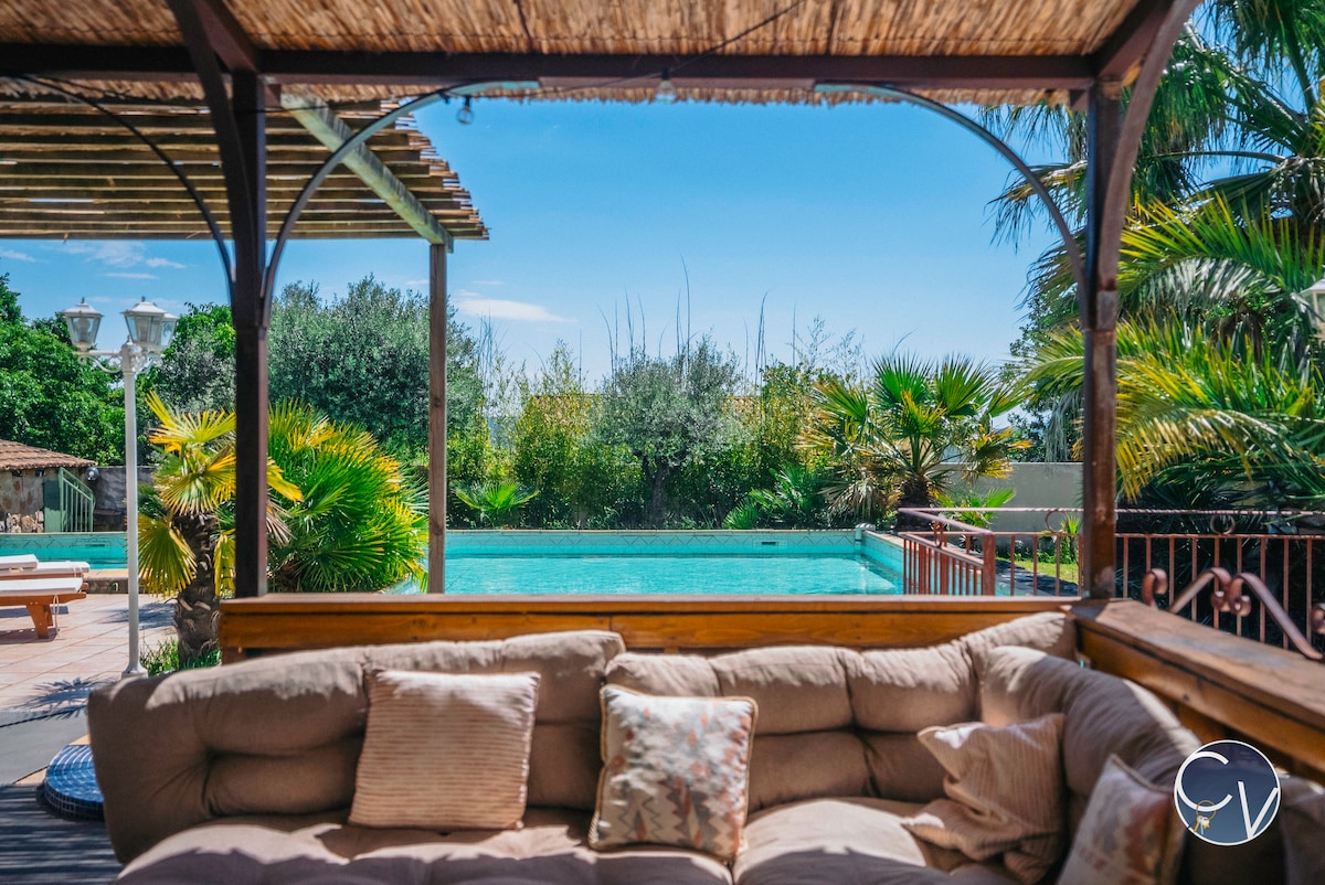 A covered outdoor lounge area, featuring comfortable seating with cushions, overlooks a spacious private pool surrounded by lush greenery. Sunlight filters through the palm trees, creating a serene atmosphere. The pool reflects the clear blue sky and adds to the inviting setting.