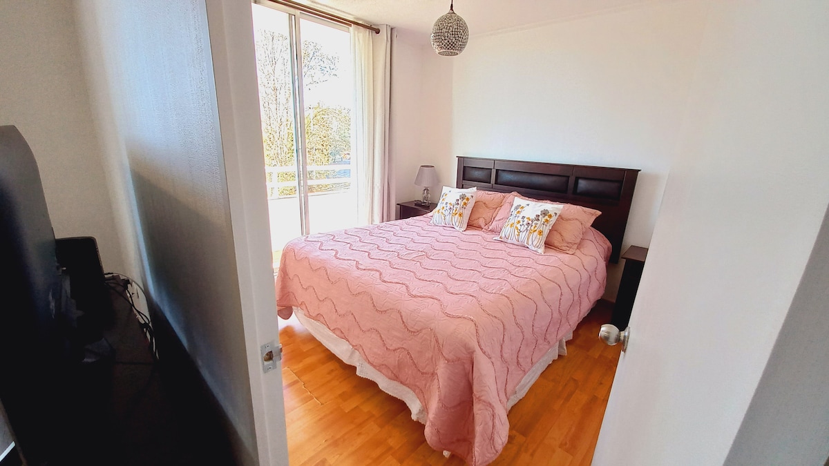 A well-lit bedroom features a spacious bed covered with a light pink quilt and accented by two decorative pillows. Natural light enters through sliding glass doors, illuminating the wooden flooring. A nightstand and a lamp are visible beside the bed, contributing to the room's functionality.