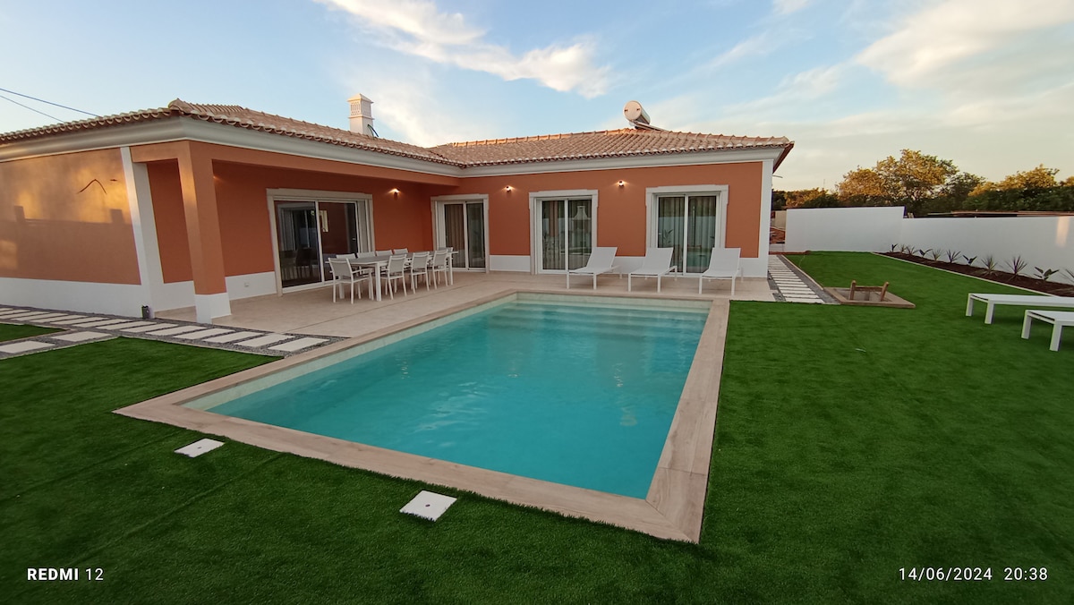 A charming villa features a private pool surrounded by lush green grass. The outdoor seating area showcases a table with several chairs. Large glass doors lead into the inviting interior, where natural light enhances the warm tones of the structure.