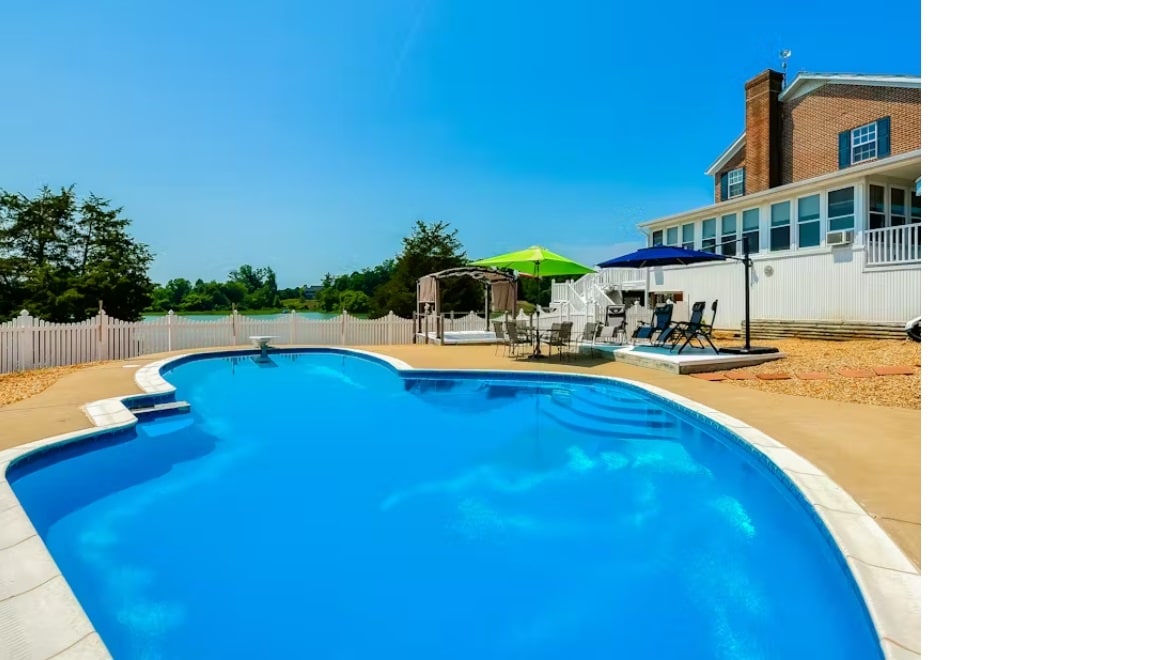 A bright blue pool sparkles under clear skies, surrounded by a patio area with several lounge chairs and shaded spaces. The home, featuring large windows, stands in the background, inviting relaxation and outdoor enjoyment.