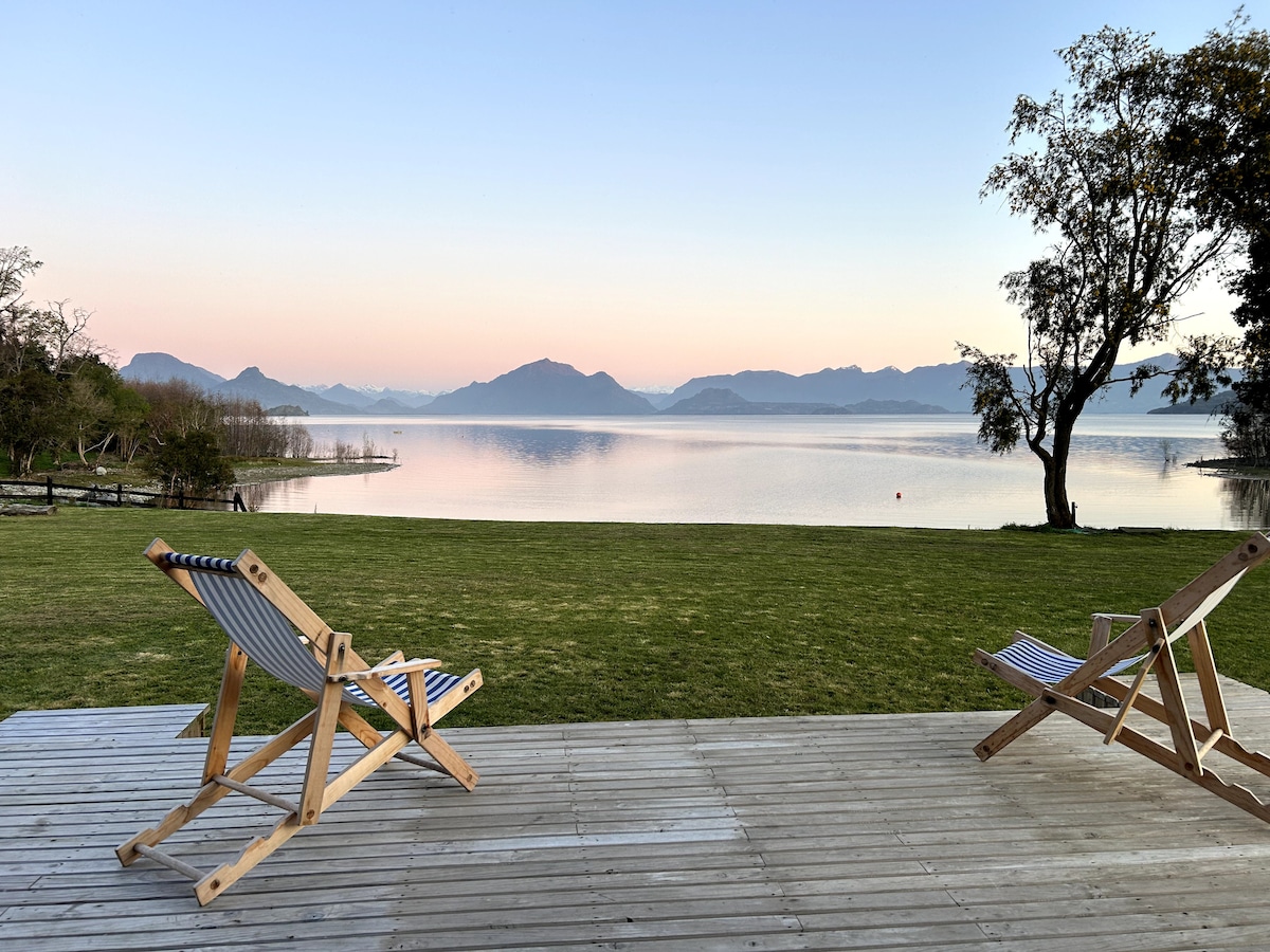 A serene outdoor space features two wooden lounge chairs positioned on a wooden deck. A vast lawn leads to a tranquil lake, reflecting the soft colors of the sunrise. Mountains are visible in the distance, creating a peaceful backdrop.