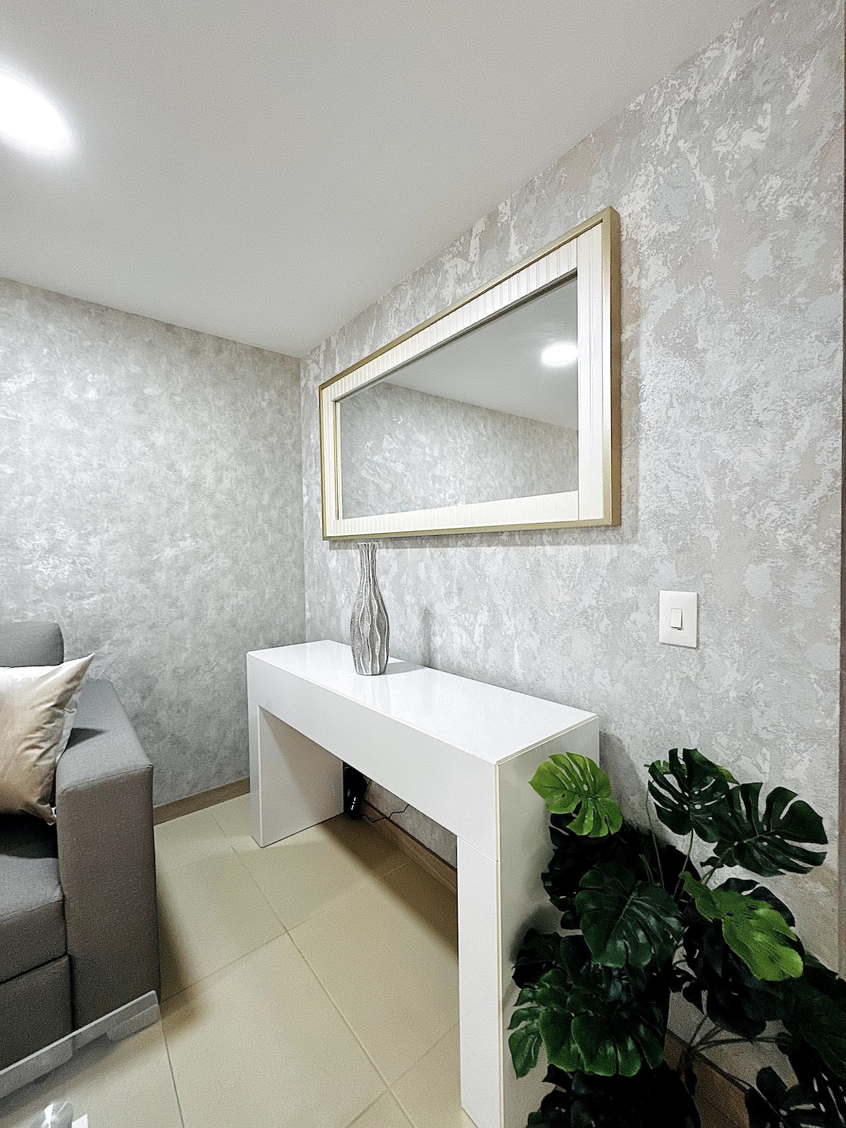 Ukrasni kutak s teksturiranim zidom, modernim bijelim stolom, elegantnom vazom i velikim zrcalom koje proširuje svjetlost prostora. Biljka biljka dodaje svježinu i ugodnu atmosferu.

