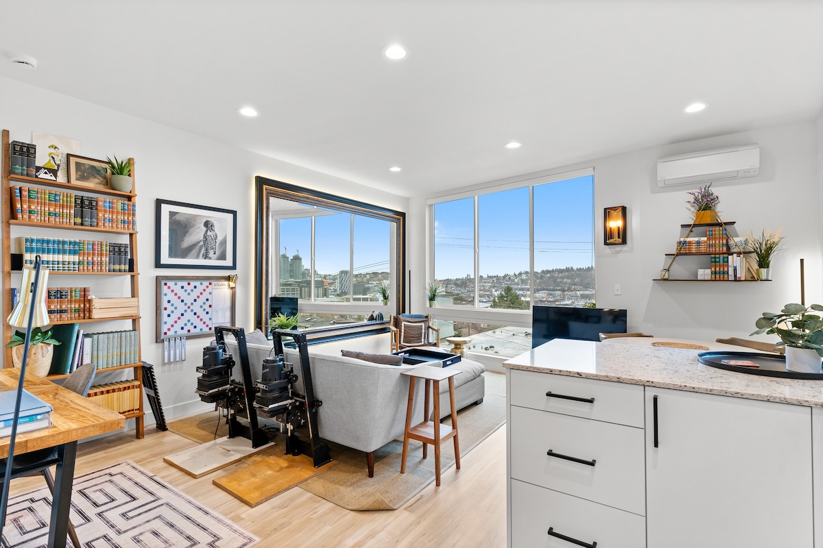 A bright and modern living area is highlighted by large windows offering views of the Seattle skyline. A comfortable sofa sits on a light wood floor, complemented by a cozy area rug. Shelves filled with books and artwork contribute to the inviting ambiance.