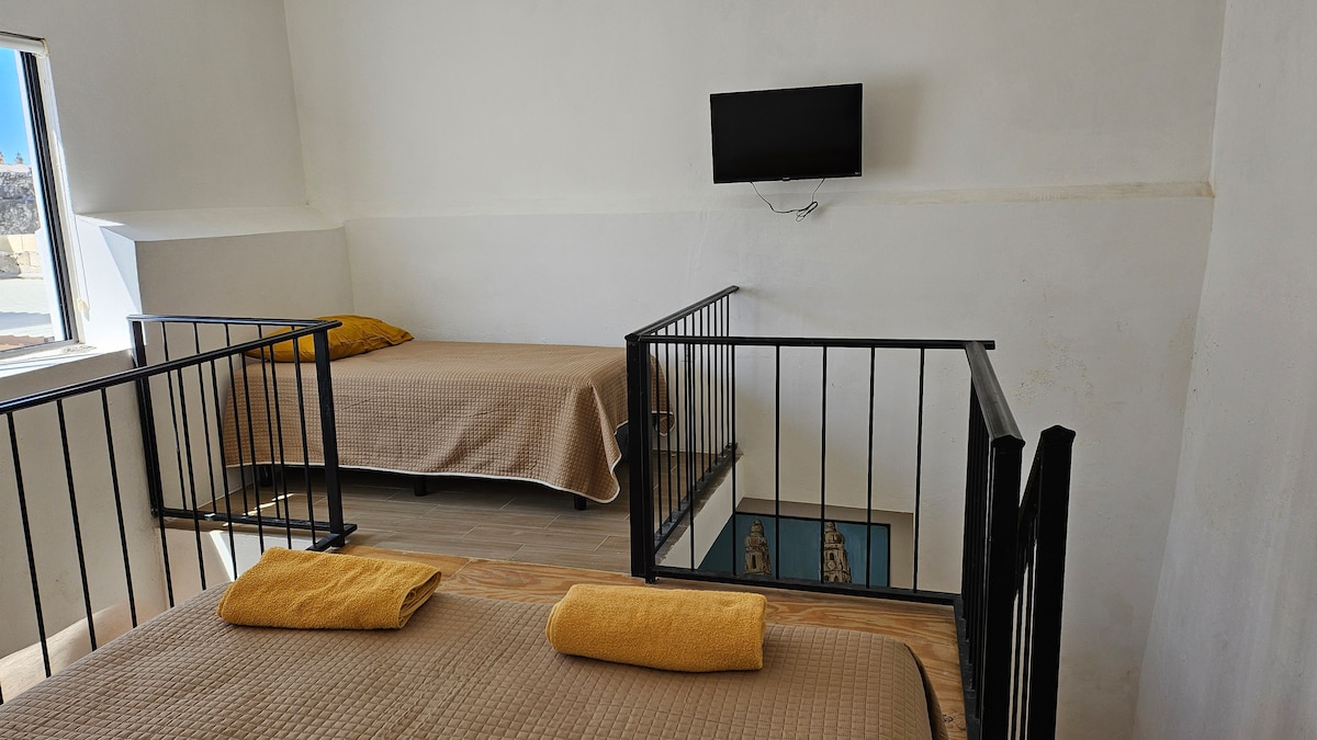 A lofted sleeping area showcases two beds, one covered with a light brown blanket and a second with a yellow accent pillow. A small television is mounted on the wall. Bright light filters in through a nearby window, contributing to the open and airy feel.