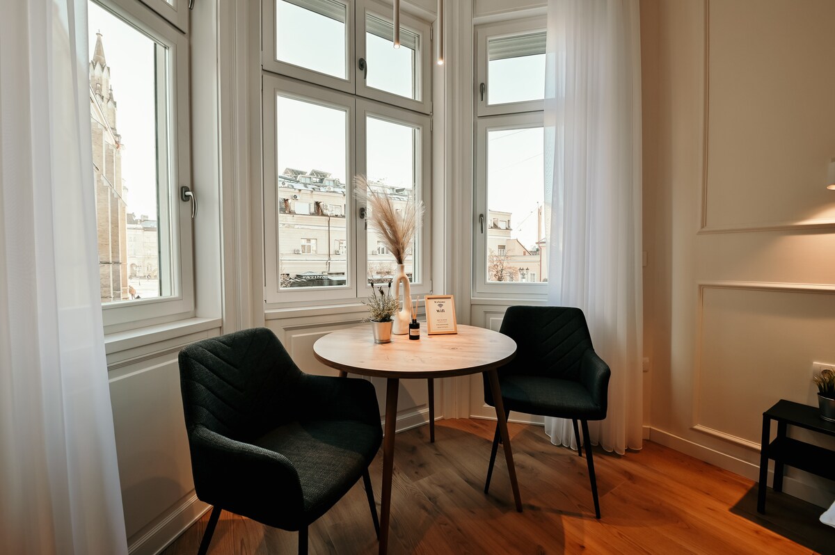 A contemporary seating area is highlighted by a round table and two dark upholstered chairs. Large windows allow natural light to fill the space, offering views of the cityscape and soft white curtains that frame the scene.