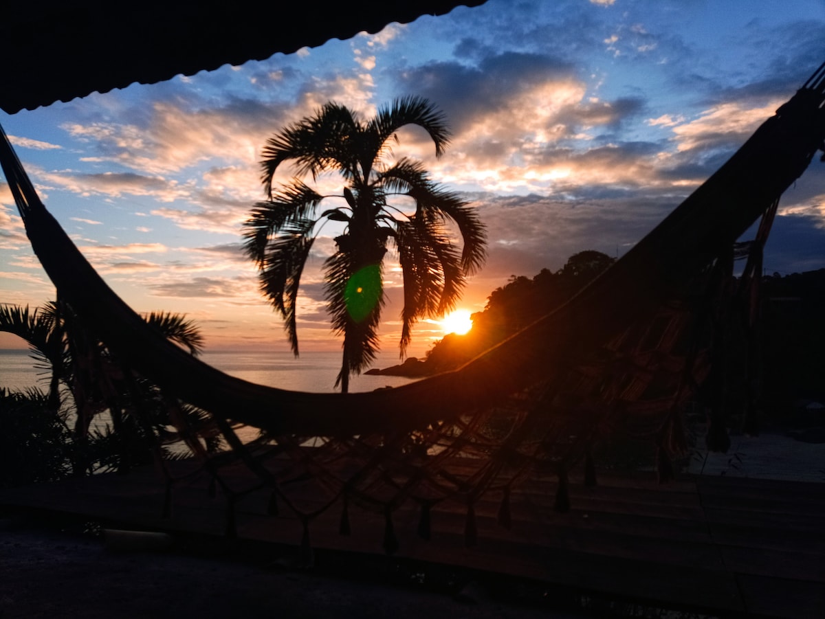A hammock is framed by palm trees in the foreground, with the sun setting on the horizon. The sky is filled with soft clouds reflecting warm hues of orange and yellow, while the sea glistens in the fading light. The tranquil scene invites relaxation.