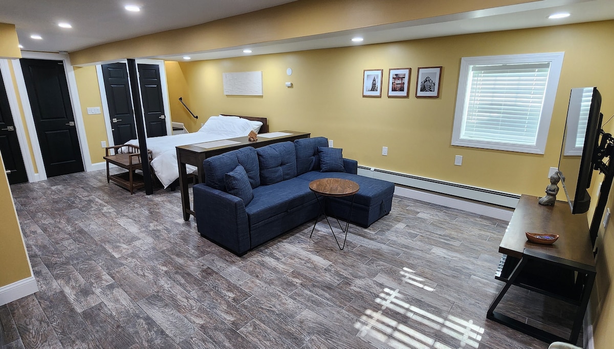The space features a stylish seating area with a dark blue sofa and a small round table. A spacious bed is positioned in the background, while black doors and framed art add character to the room. Natural light enters through a window, illuminating the area.