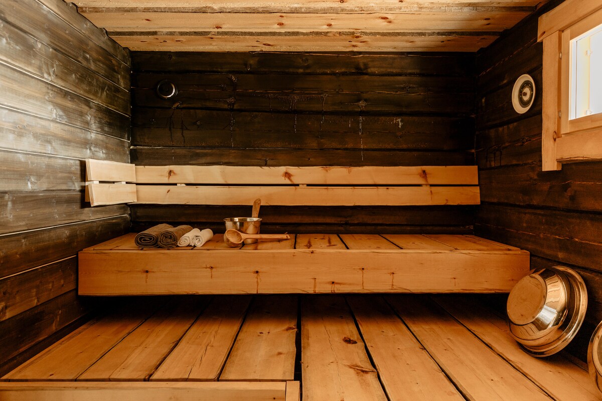A traditional wood-heated sauna is showcased with wooden benches along the walls. Natural wood tones create a warm environment, complemented by steam buckets and towels neatly arranged on the floor, highlighting the sauna's practical features.
