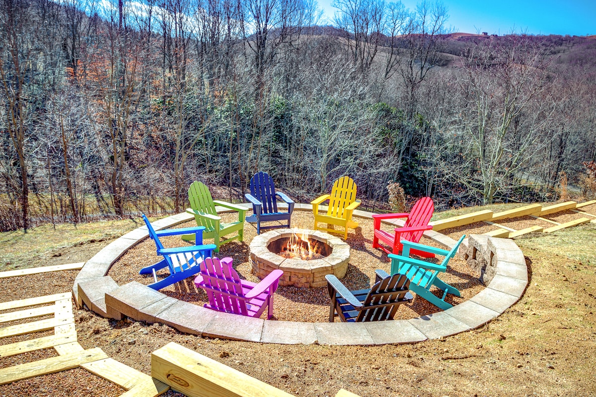 An outdoor fire pit area features a circular arrangement of colorful Adirondack chairs surrounding a central stone fire pit. The setting is surrounded by trees, and the open landscape provides a scenic view of the distant hills on a clear day.