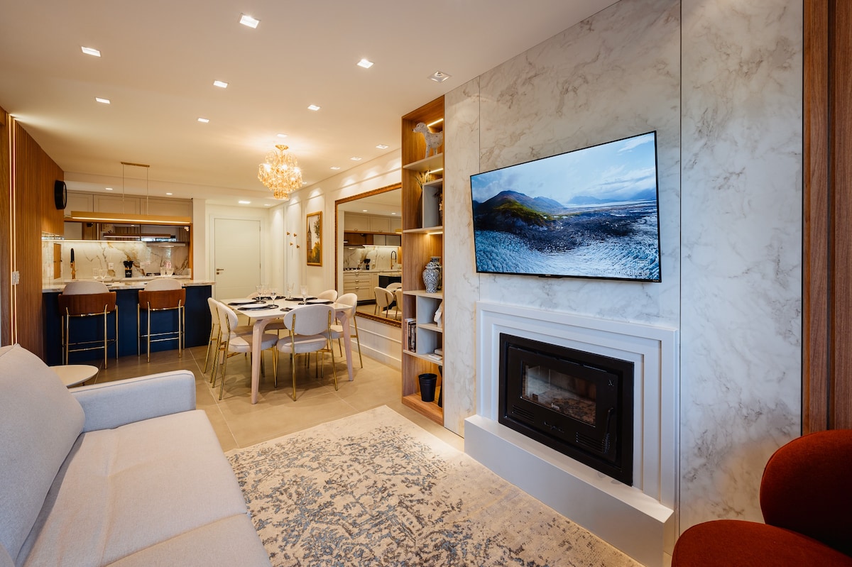 A modern living area features a light-colored sofa facing a fireplace and a large wall-mounted television. An elegant dining space is visible in the background, alongside an open kitchen with a breakfast bar. Soft lighting enhances the welcoming ambiance.