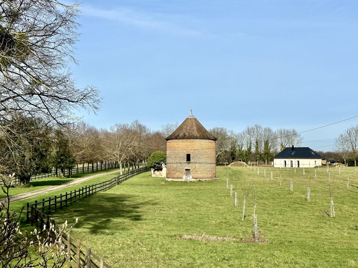 A historic round building with a thatched roof is visible in an open grassy area, surrounded by trees and a wooden fence. A pathway leads to the structure, while a smaller house is seen in the background, amidst a tranquil rural landscape.