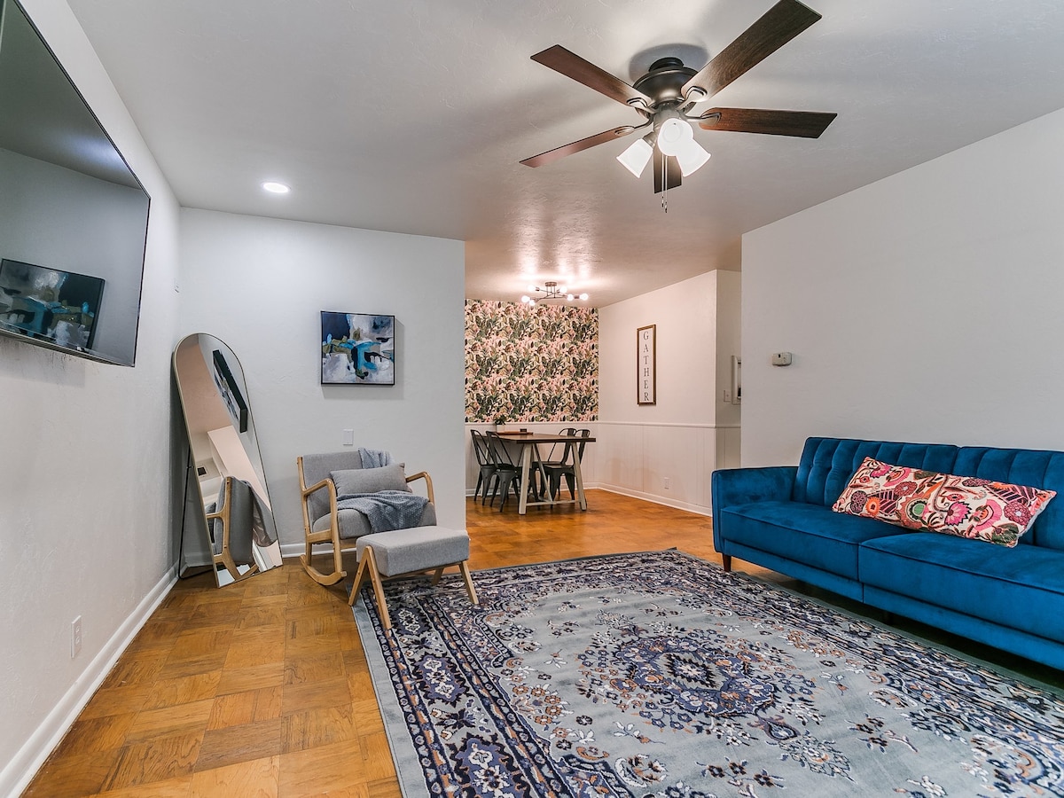 A living area features a deep blue couch, a stylish mirror, and a rocking chair. A decorative area rug provides comfort underfoot, while a ceiling fan offers ventilation. The adjoining dining area showcases floral wallpaper and a spacious table, enhancing the inviting ambiance.