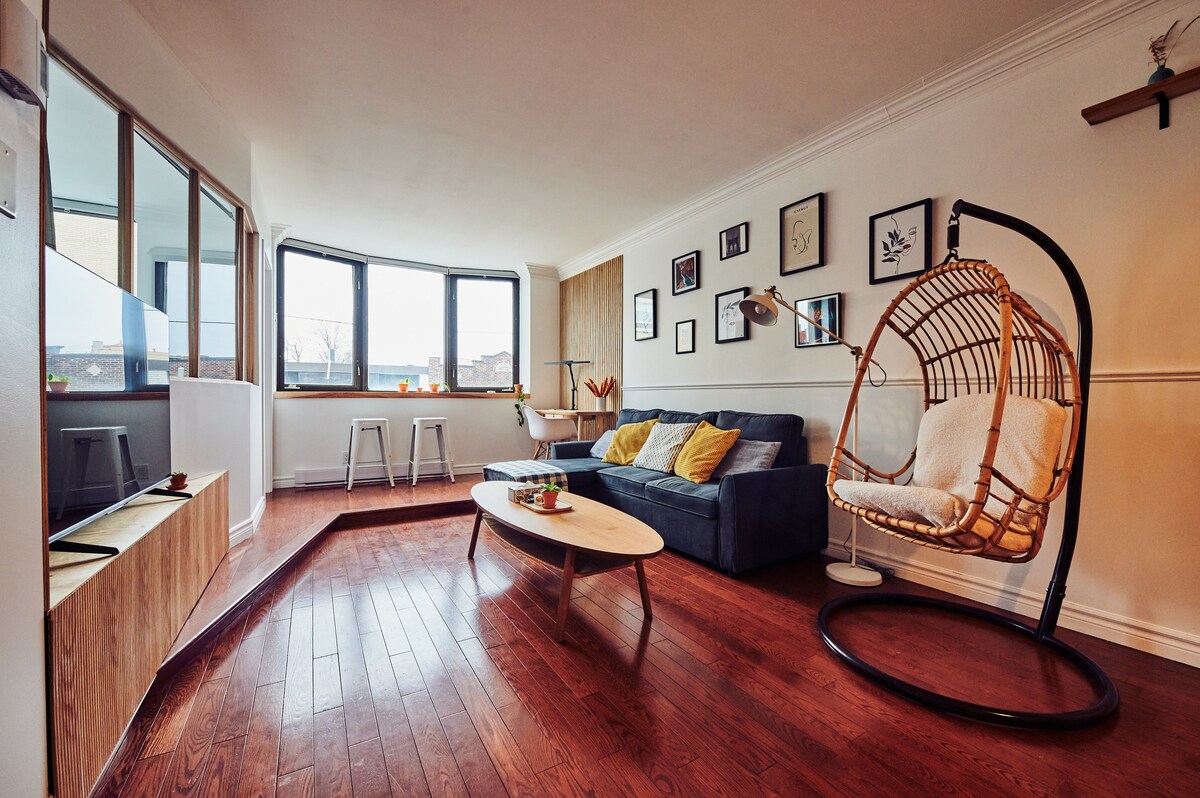 A spacious living area features large windows allowing natural light to flow in. A comfortable blue sofa is complemented by cheerful yellow cushions. A modern coffee table sits on polished wooden flooring. A unique hanging chair adds a stylish touch, while framed artworks decorate the walls.