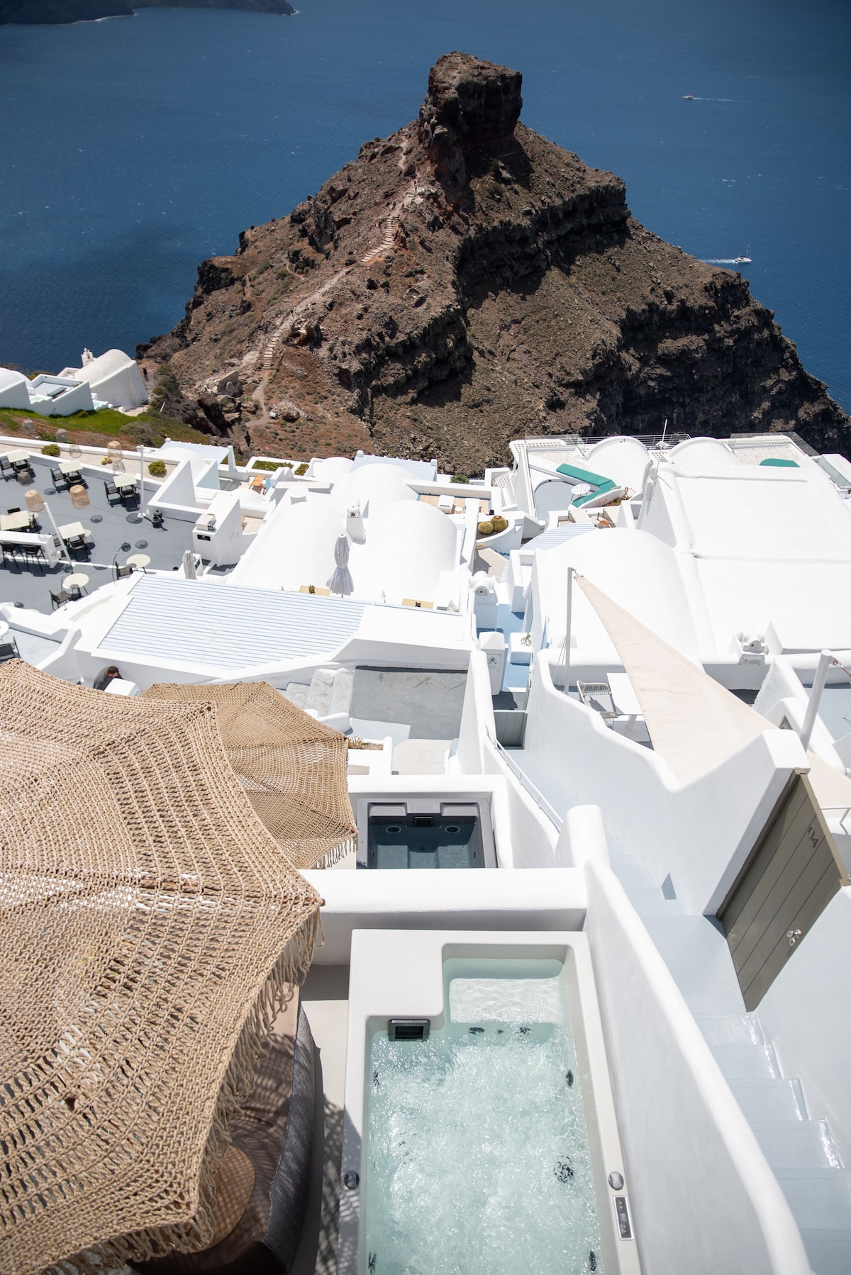 A view from above captures a private hot tub on a terrace, surrounded by whitewashed buildings. Below, rocky cliffs descend into the deep blue sea. The sunny landscape highlights the rugged terrain and creates a serene coastal atmosphere.