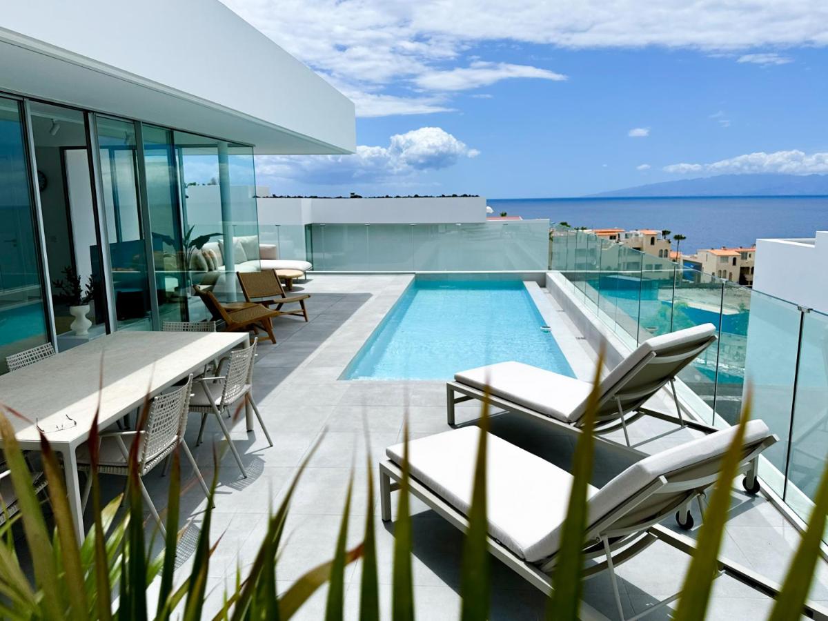 An outdoor terrace features a sleek swimming pool surrounded by lounge chairs, offering a view of the ocean. A dining table is positioned nearby, complemented by tall glass railings that enhance the open atmosphere. Lush greenery adds a natural touch to the space.