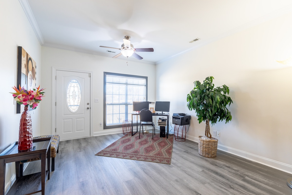 A welcoming living space features a ceiling fan and a natural light-filled area with a desk and chairs set up for work. A decorative plant adds a touch of greenery, while colorful artwork and a red-patterned rug enhance the inviting atmosphere.