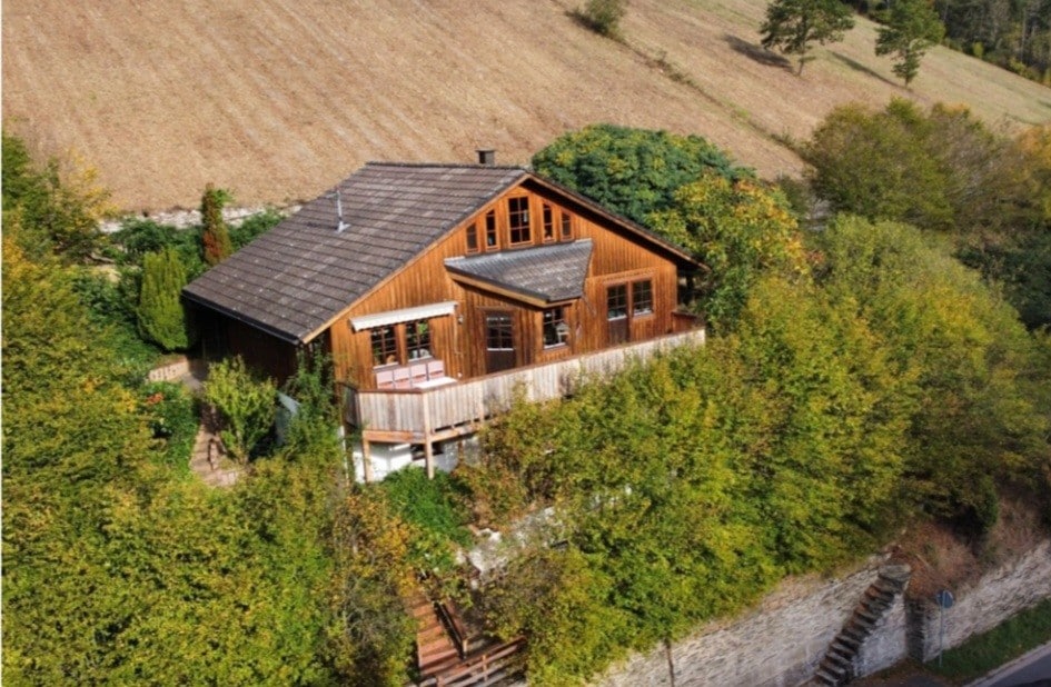 A wooden house is nestled among greenery on a hillside, featuring a large balcony that overlooks the landscape. The exterior showcases large windows and a sloped roof, blending harmoniously with the natural environment.