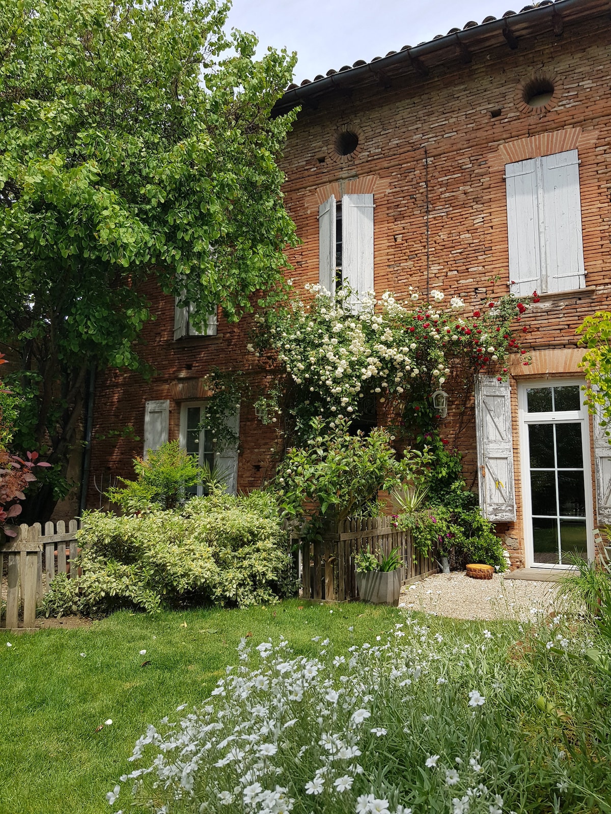 The exterior of a brick house is framed by lush greenery and flowering vines. White shutters are partially opened, revealing a glimpse of the interior. A small fenced garden features a mix of plants and flowers, contributing to the inviting atmosphere of the property.