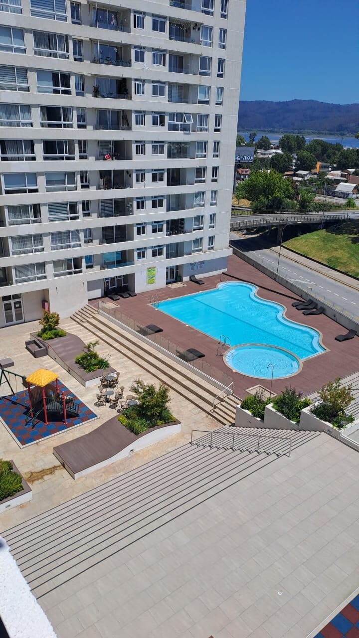 An overview of the outdoor pool area is shown, featuring two swimming pools surrounded by lounge chairs. Play equipment is visible to the side, along with landscaped areas adding greenery. The multi-story building stands adjacent, providing a modern residential backdrop.