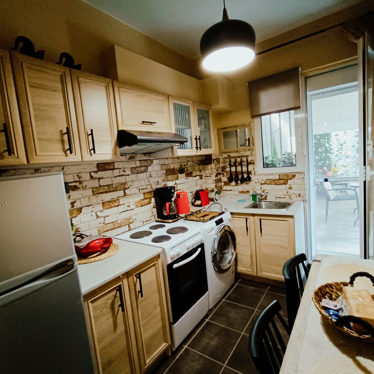A well-equipped kitchen is depicted, featuring light wooden cabinets, a stove, and an oven. A washing machine is positioned next to the refrigerator. Counter space offers appliances like a coffee maker and kettle, while a door leads to an outdoor area, enhancing the openness of the space.