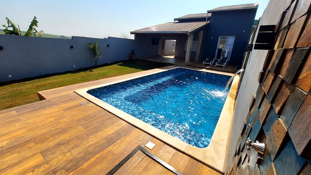 A spacious outdoor area features a large swimming pool with clear blue water and a deck made of wood. A gentle waterfall is visible flowing into the pool. Sun loungers are positioned nearby, and the surrounding grass is well maintained.
