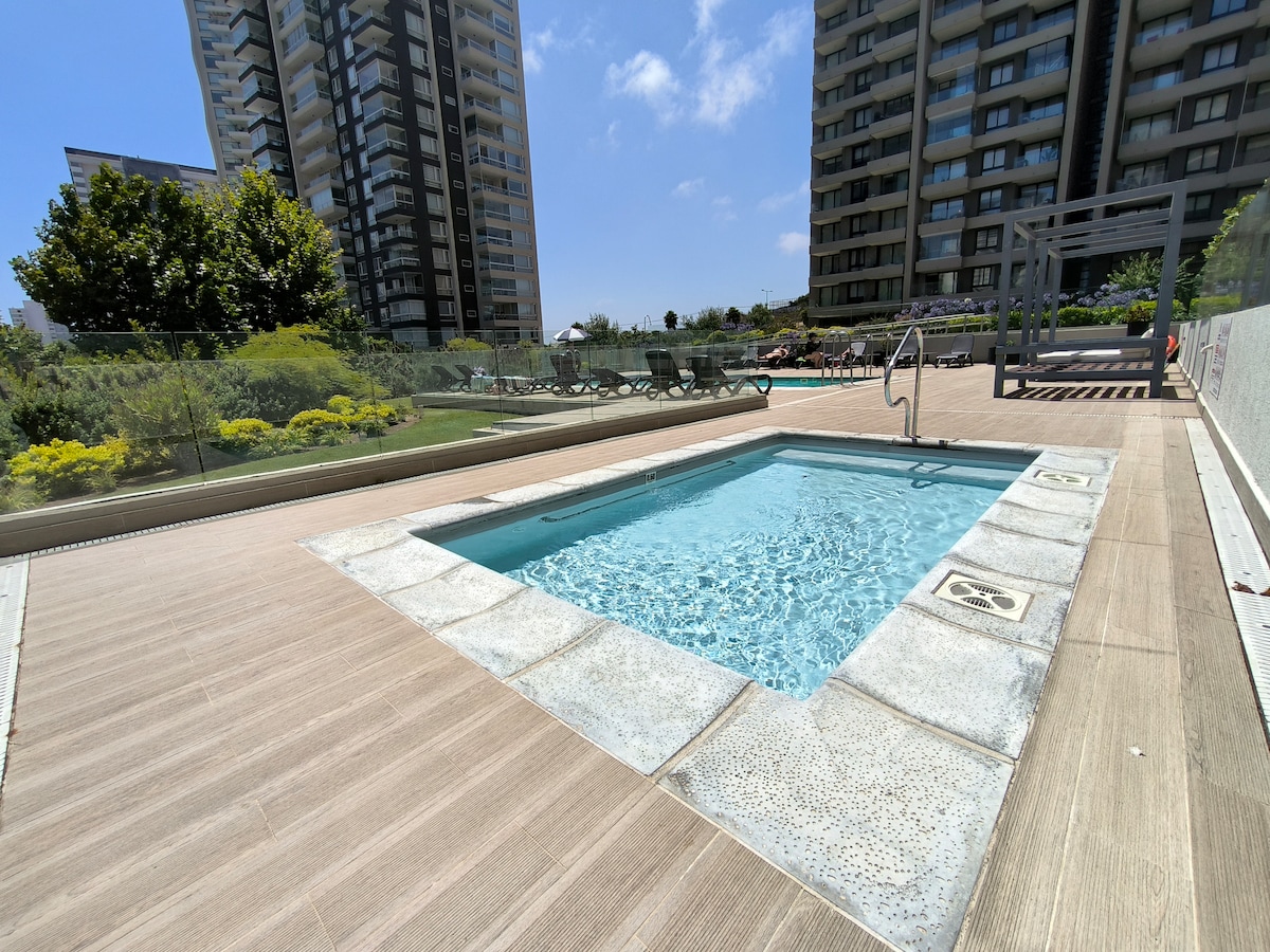 A private jacuzzi is seen on a wooden deck, surrounded by lush greenery. Sunlight reflects off the water, with lounge chairs positioned nearby and modern apartment buildings in the background. The area is designed for relaxation and leisure.