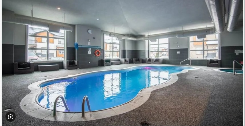 An indoor pool area features a uniquely shaped pool with clear blue water. Comfortable seating is arranged along the edges, and windows allow natural light to brighten the space. A hot tub is visible in the corner, enhancing the recreational atmosphere.