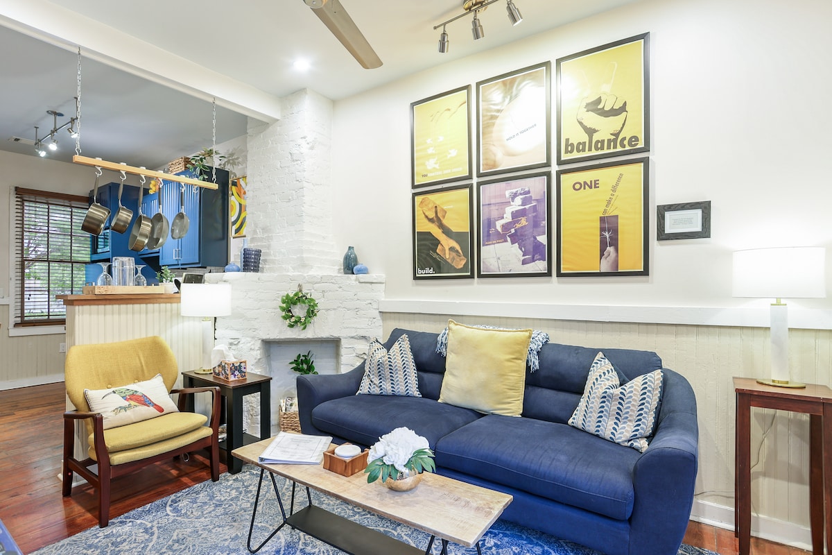 The living area features a dark blue sofa paired with a wooden coffee table and an accent chair. Bright artwork adorns the wall above, while recessed lighting showcases the space. A glimpse of the kitchen is visible in the background, showcasing blue cabinetry and hanging pots.