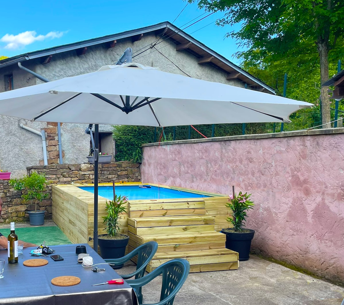 A small pool is surrounded by wooden decking, providing a cozy outdoor space. An umbrella offers shade over a dining area with a round table and chairs. Potted plants add greenery, while a stone wall and trees complete the tranquil atmosphere.
